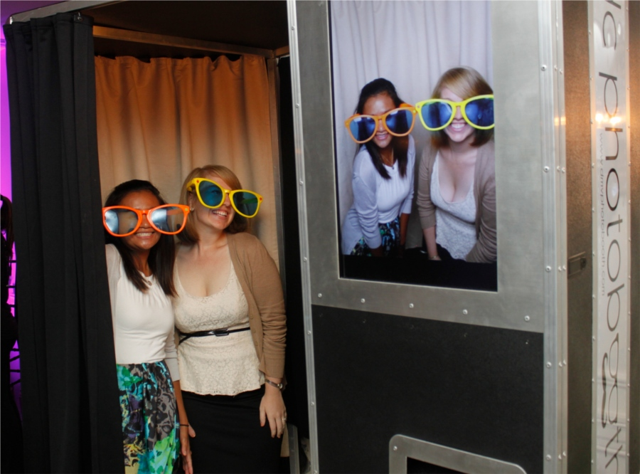 Two friends wearing oversized sunglasses having fun inside the Raleigh Photobooths enclosed photo booth
